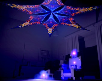 Puede incluir: Un gran tejido en forma de estrella multicolor que cuelga del techo. El tejido tiene un patrón psicodélico con colores azules, naranjas y amarillos. El tejido está colgado en una habitación con paredes y cortinas blancas.