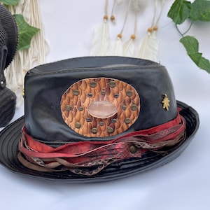 May include: Black fedora hat with a decorative oval patch featuring a light brown snakeskin pattern, copper studs, and a central pink stone. The hat has a red and brown ribbon band and a gold-colored leaf pin.