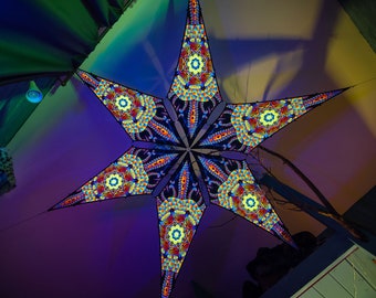 Puede incluir: Una gran decoración colgante en forma de estrella multicolor con intrincados patrones. La estrella está hecha de tela y tiene un contorno negro. La estrella está colgada del techo y está iluminada por luces de colores.