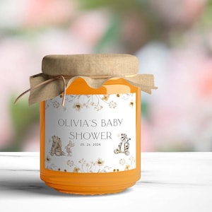 May include: A glass jar filled with honey, decorated with a white label featuring Winnie the Pooh and friends. The label reads "Olivia's Baby Shower 05.26.2024".