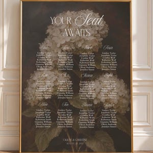 May include: A rectangular wedding seating chart sign in a gold frame featuring a dark background with white hydrangea flowers. The text reads YOUR SEAT AWAITS, lists tables one through twelve with names, and OLIVER & CHRISTINE AUGUST 22, 2027.