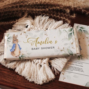May include: A white chocolate bar wrapper with a watercolor design of green leaves and gold glitter. The wrapper features a cartoon illustration of Peter Rabbit and the text "Amelia's Baby Shower".