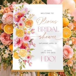 May include: A white sign on a gold easel, announcing "Olivia's Bridal Shower." The sign is decorated with watercolor flowers and orange slices. The text includes the date, and the phrase "I Do!" surrounded by floral arrangements and balloons.