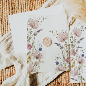 Puede incluir: Un sobre blanco con un diseño floral y un sello de cera. El sobre está sobre una estera tejida con un paño blanco drapeado sobre él.