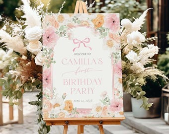 May include: A rectangular birthday party sign on a wooden easel featuring a pink bow graphic, floral illustrations in shades of pink, orange, and white, and text reading Welcome to Camilla's First Birthday Party June 22, 2025.