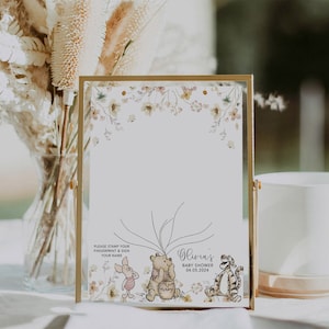 May include: A Winnie the Pooh themed baby shower guest book with a floral border. The guest book has a white background with a space for guests to sign their names and leave fingerprints. The text on the guest book reads "Please stamp your fingerprint & sign your name" and "Olivia's Baby Shower 04.05.2024".