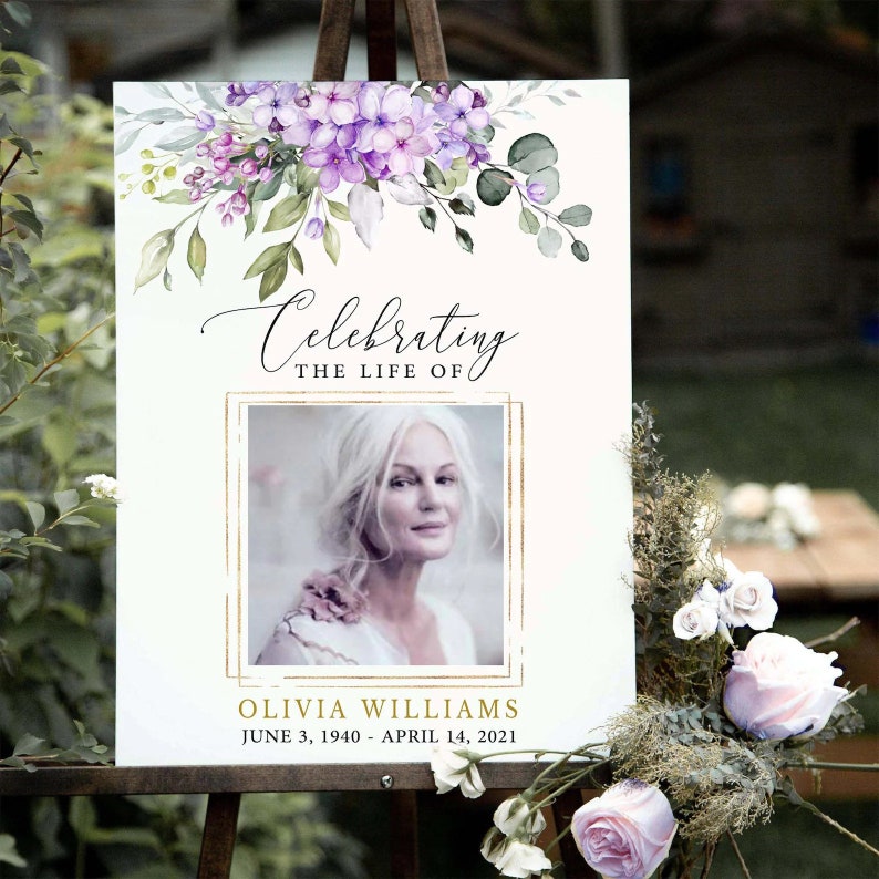 May include: A white memorial sign with a gold frame and a floral design at the top. The sign reads "Celebrating the Life of" and includes a photo of a woman, the name "Olivia Williams", and the dates "June 3, 1940 - April 14, 2021".