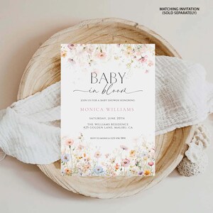 May include: A baby shower invitation with floral details. The invitation says "BABY in bloom" and includes details for Monica Williams' baby shower on June 20th at The Williams Residence in Malibu, CA. The invitation is on a wooden plate with white fabric.