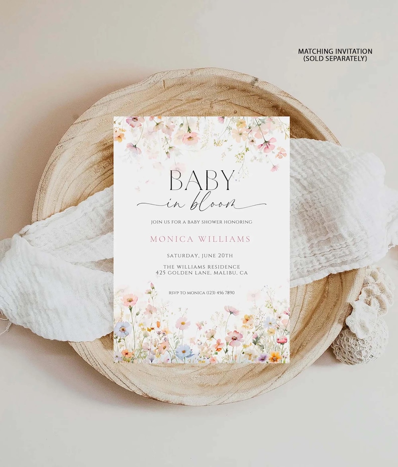May include: A baby shower invitation with floral details. The invitation says "BABY in bloom" and includes details for Monica Williams' baby shower on June 20th at The Williams Residence in Malibu, CA. The invitation is on a wooden plate with white fabric.