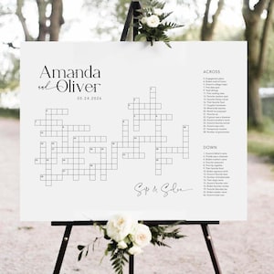May include: A white wedding sign with a crossword puzzle design. The sign displays the names "Amanda and Oliver" and the date "05.24.2026". The sign is propped up on a black easel and decorated with white flowers.