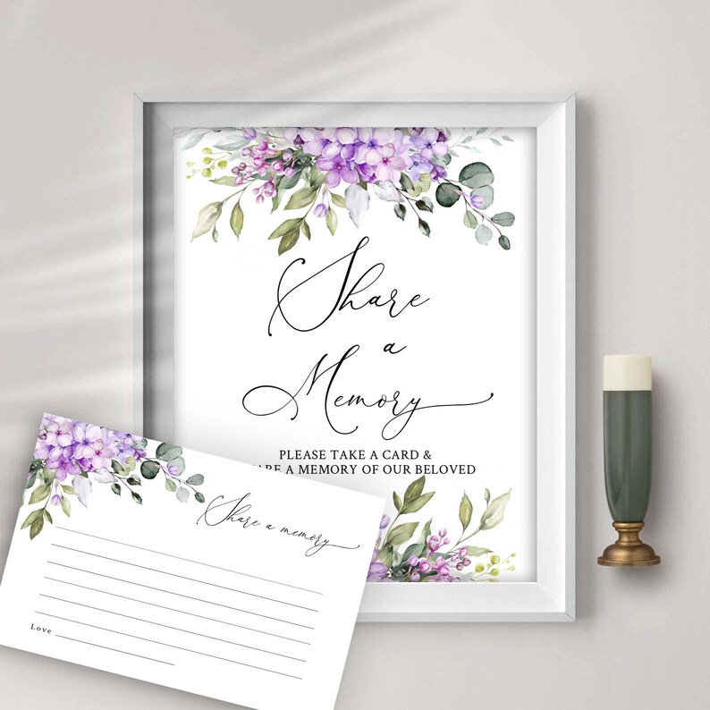 May include: A white sign with purple flowers and greenery at the top. The sign says "Share a Memory" and "Please take a card & write a memory of our beloved". There is a smaller white card with purple flowers and greenery at the top that says "Share a memory" and has lines for writing.