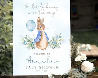 May include: A rectangular baby shower sign featuring a watercolor illustration of a brown rabbit in a blue jacket holding a light blue balloon. The text reads A little bunny is on the way! Welcome to Amanda's Baby Shower May 17, 2021.