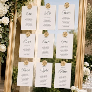 May include: A large gold-framed mirror displaying nine white rectangular table seating cards arranged in three rows. Each card features a gold wax seal at the top, the word TABLE followed by the number in script, and a list of six names.