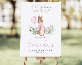 May include: A white rectangular sign on a wooden easel featuring a watercolor illustration of a brown rabbit in a pink jacket holding a pink balloon, surrounded by greenery. Text reads: A little bunny is on the way! WELCOME TO Amanda's BABY SHOWER APRIL 17, 2025.