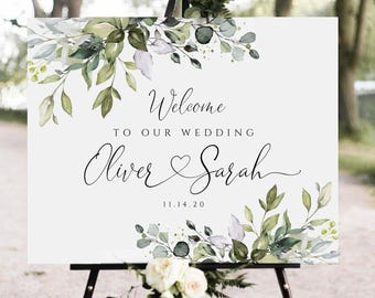 May include: A rectangular wedding welcome sign featuring green eucalyptus leaves and white floral accents. The text reads Welcome to our wedding, Oliver and Sarah, and the date 11.14.20 in black script and serif fonts on a white background.