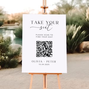May include: A white sign on a wooden easel with the text "TAKE YOUR seat" in elegant script. Below, it reads "PLEASE SCAN TO FIND YOUR SEAT" with a QR code. The names "OLIVIA + PETER" and the date "10.04.2025" are at the bottom.