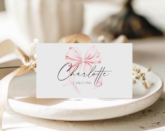 May include: A rectangular white place card featuring a soft pink watercolor illustration of a bow centered above the name Charlotte in black cursive script, with the text TABLE ONE printed in smaller black capital letters below the name.