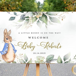 May include: A white welcome sign with a watercolour illustration of a bunny rabbit, green leaves, and gold accents. The text reads "A little bunny is on the way! Welcome Baby Roberts 10.14.2019".