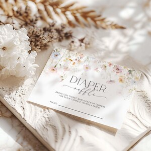 May include: A white "Diaper Raffle" ticket with floral detailing. The card displays the words "Diaper Raffle" in a stylish script, with instructions and a space for a name. The design incorporates soft watercolor flowers in pastel shades.