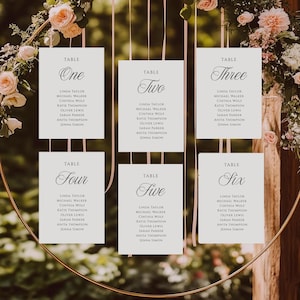 Puede incluir: Seis tarjetas rectangulares blancas de distribución de mesas de boda con texto en negro, colgadas de un marco circular dorado. Cada tarjeta muestra un número de mesa del uno al seis en letra cursiva, seguido de una lista de nombres en letra sans-serif.