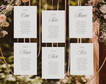 May include: Six white rectangular wedding seating chart cards with black text hanging from a circular gold frame. Each card displays a table number from one to six in a script font, followed by a list of names in a simple sans-serif typeface.