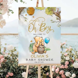 May include: A Winnie the Pooh themed baby shower sign with a blue and white watercolor background. The sign features Winnie the Pooh holding a pot of honey and a blue balloon. The text on the sign reads "Oh Boy" in gold script, "Welcome to Olivia's Baby Shower" and "06.27.25".