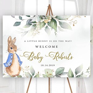 May include: A welcome sign for a baby shower with a watercolour illustration of a bunny rabbit wearing a blue jacket. The sign says "A little bunny is on the way! Welcome Baby Roberts 10.14.2019".
