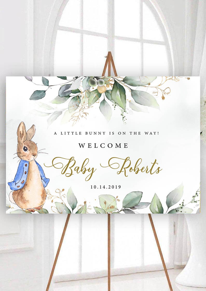 May include: A welcome sign for a baby shower with a watercolour illustration of a bunny rabbit wearing a blue jacket. The sign says "A little bunny is on the way! Welcome Baby Roberts 10.14.2019".