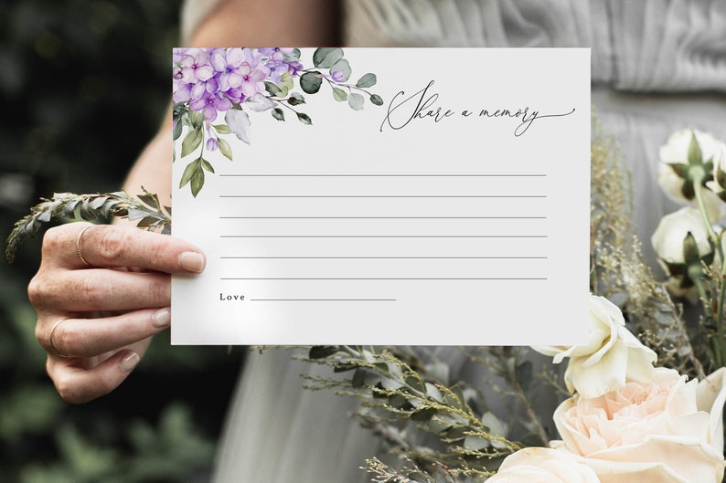 May include: A white card with a floral design in shades of purple and green. The card has the text "Share a memory" written in cursive. There is a blank space for writing and a line for a signature.