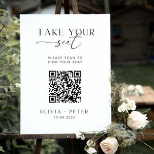 May include: A white sign on a wooden easel with the text "TAKE YOUR seat". Below, it reads "PLEASE SCAN TO FIND YOUR SEAT" with a QR code. The sign is decorated with flowers and the names Olivia + Peter and the date 10.04.2025.