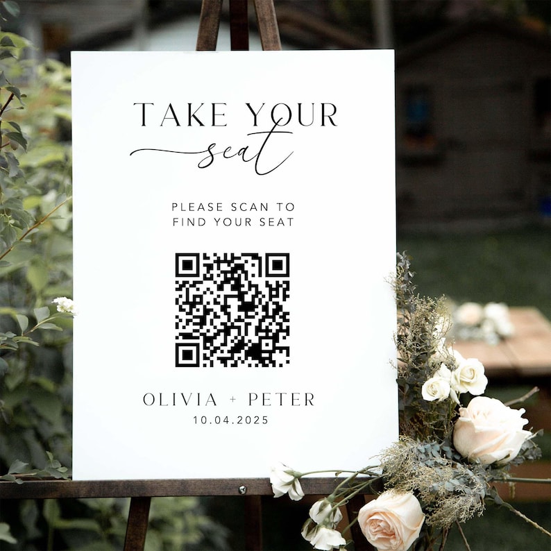 May include: A white sign on a wooden easel with the text "TAKE YOUR seat". Below, it reads "PLEASE SCAN TO FIND YOUR SEAT" with a QR code. The sign is decorated with flowers and the names Olivia + Peter and the date 10.04.2025.