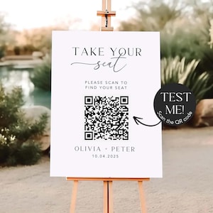May include: A white sign on a wooden easel with the text "TAKE YOUR seat" in elegant script. Below, it reads "PLEASE SCAN TO FIND YOUR SEAT" with a QR code. The sign also includes the names "OLIVIA + PETER" and the date "10.04.2025". A black circle with the text "TEST ME! Scan the QR code" is on the right.