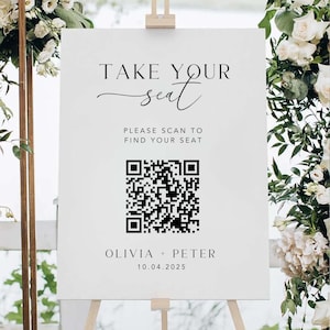 May include: A white wedding sign with the text "TAKE YOUR seat" and "PLEASE SCAN TO FIND YOUR SEAT". A QR code is below the text, followed by "OLIVIA + PETER" and the date "10.04.2025". The sign is on an easel.