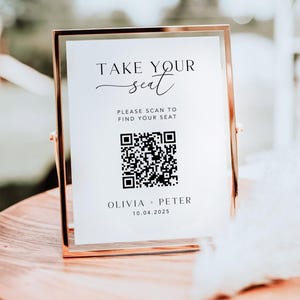 May include: A gold-framed sign with the text "TAKE YOUR seat" and a QR code. Below the QR code are the names "OLIVIA + PETER" and the date "10.04.2025". The sign is displayed on a wooden surface, likely for a wedding or event.