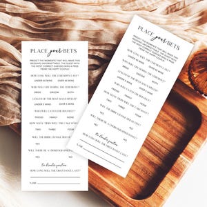 May include: Two white wedding game cards with the text "Place Your Bets" are displayed on a wooden tray. The cards have a list of questions about the wedding. The background includes a beige fabric and decorative elements.