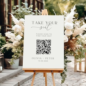 May include: A cream-colored sign on a wooden easel with the text "TAKE YOUR seat". Below is a QR code and the text "PLEASE SCAN TO FIND YOUR SEAT". The bottom of the sign reads "OLIVIA + PETER 10.04.2025". The sign is surrounded by white flowers and greenery.