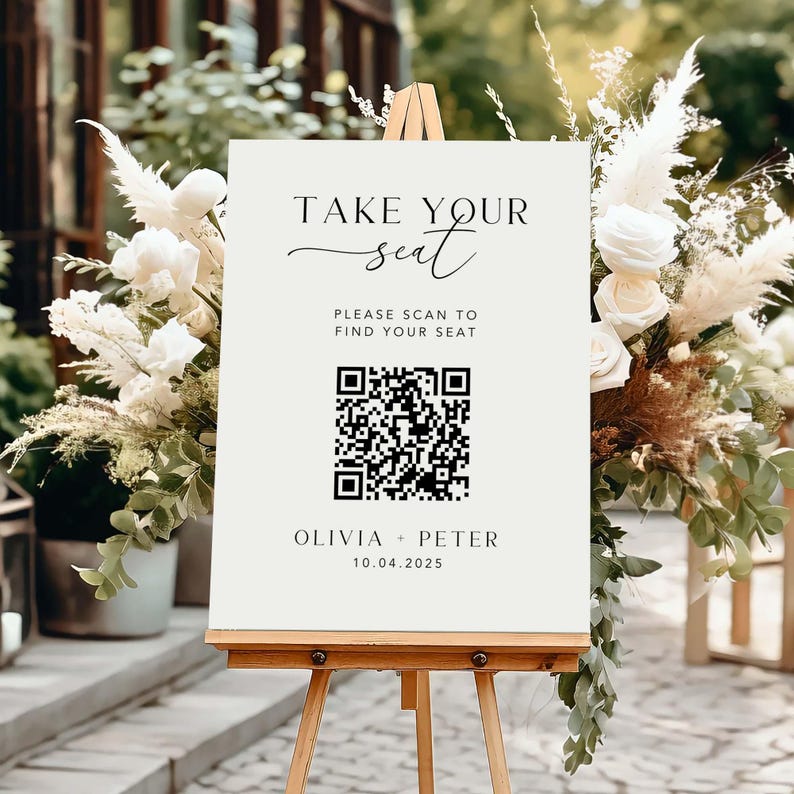 May include: A cream-colored sign on a wooden easel with the text "TAKE YOUR seat". Below is a QR code and the text "PLEASE SCAN TO FIND YOUR SEAT". The bottom of the sign reads "OLIVIA + PETER 10.04.2025". The sign is surrounded by white flowers and greenery.