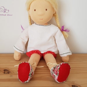 May include: A handmade doll with blonde hair, wearing a white long-sleeved shirt, red and white polka dot shorts, and red and floral print shoes.