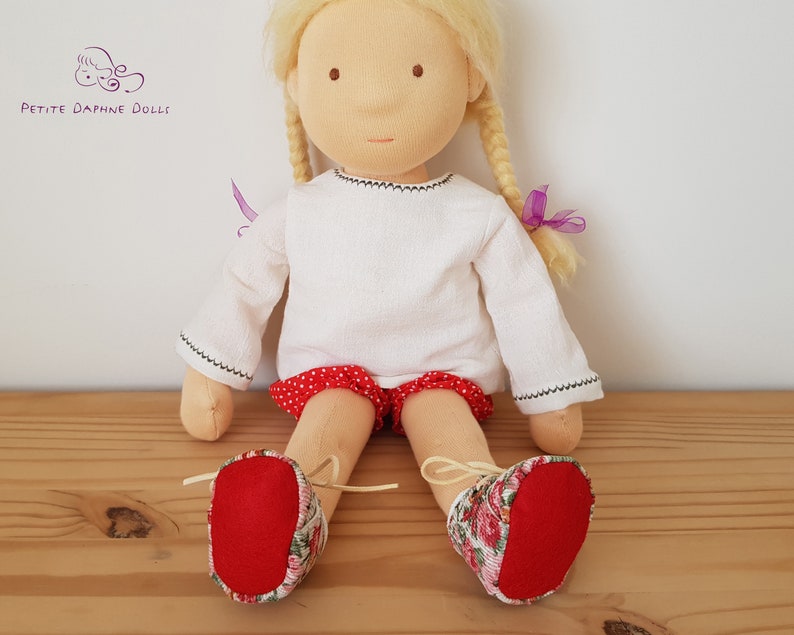 May include: A handmade doll with blonde hair, wearing a white long-sleeved shirt, red and white polka dot shorts, and red and floral print shoes.
