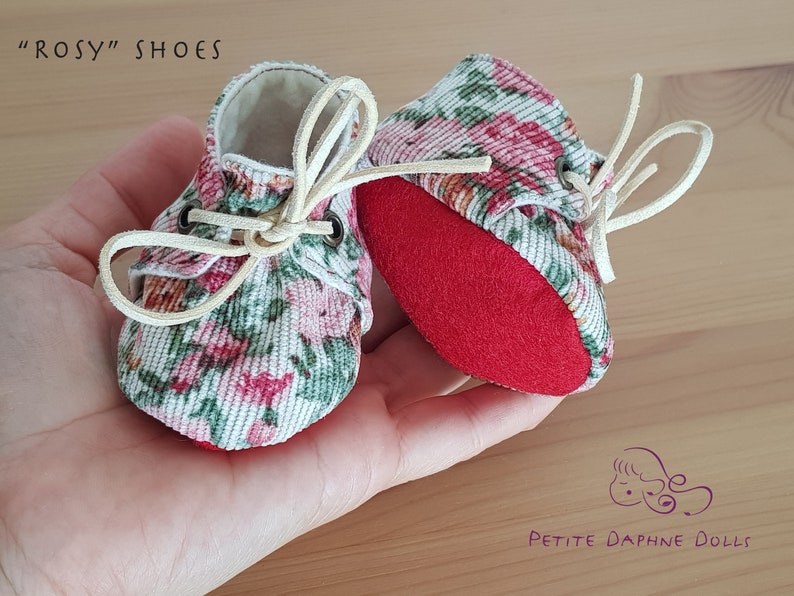May include: A pair of miniature floral print shoes with red soles and white laces. The shoes are tied with a bow and the text "ROSY" SHOES is visible in the image.