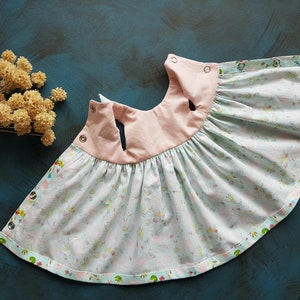 Puede incluir: Un vestido de beb&eacute; azul claro y rosa con estampado floral y cierres a presi&oacute;n. El vestido tiene una falda fruncida y un corpi&ntilde;o rosa.