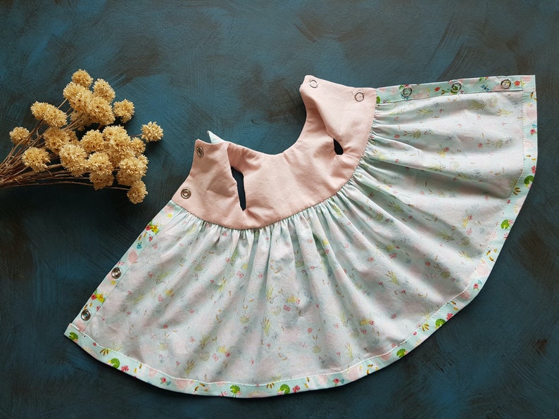 Puede incluir: Un vestido de beb&eacute; azul claro y rosa con estampado floral y cierres a presi&oacute;n. El vestido tiene una falda fruncida y un corpi&ntilde;o rosa.