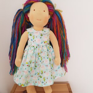 Puede incluir: Una mu&ntilde;eca hecha a mano con el pelo largo de hilo de arco&iacute;ris, vistiendo un vestido azul claro con un estampado floral. La mu&ntilde;eca tiene un cuello blanco y est&aacute; de pie sobre una superficie de madera.