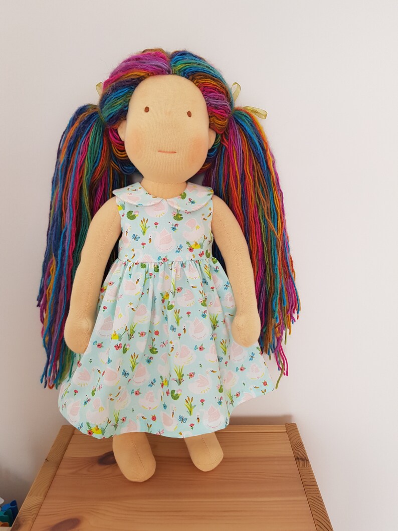 Puede incluir: Una mu&ntilde;eca hecha a mano con el pelo largo de hilo de arco&iacute;ris, vistiendo un vestido azul claro con un estampado floral. La mu&ntilde;eca tiene un cuello blanco y est&aacute; de pie sobre una superficie de madera.