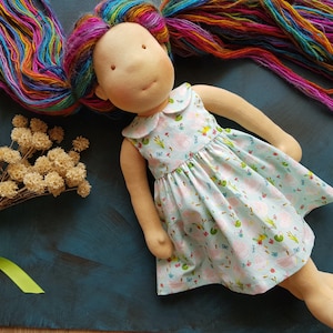 Puede incluir: Una mu&ntilde;eca hecha a mano con cabello largo de hilo arco&iacute;ris que lleva un vestido azul claro con estampado floral. El vestido tiene un cuello blanco y es sin mangas. La mu&ntilde;eca est&aacute; tumbada sobre una superficie azul.