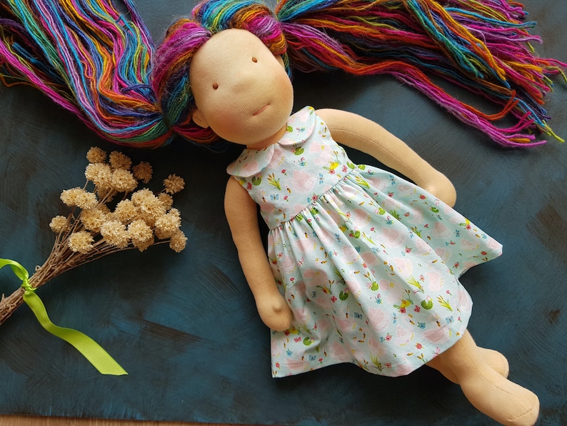 Puede incluir: Una mu&ntilde;eca hecha a mano con cabello largo de hilo arco&iacute;ris que lleva un vestido azul claro con estampado floral. El vestido tiene un cuello blanco y es sin mangas. La mu&ntilde;eca est&aacute; tumbada sobre una superficie azul.