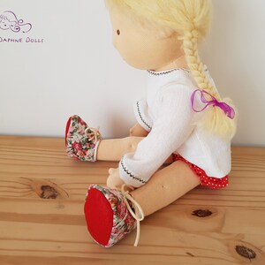 May include: A handmade doll with blonde hair in a braid tied with a purple ribbon. The doll is wearing a white long-sleeved shirt, a red and white polka dot skirt, and red shoes with floral print.