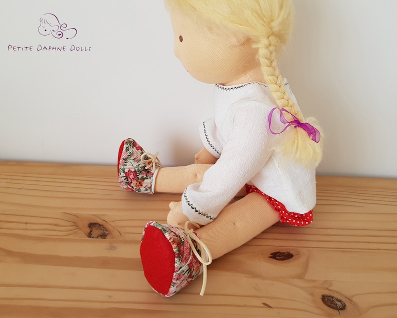May include: A handmade doll with blonde hair in a braid tied with a purple ribbon. The doll is wearing a white long-sleeved shirt, a red and white polka dot skirt, and red shoes with floral print.
