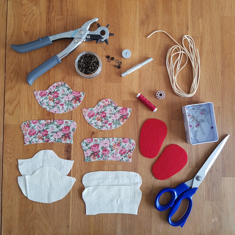 May include: A set of fabric pieces for crafting, including floral patterned fabric, plain white fabric, and red felt. The image also shows tools for crafting, such as a hole punch, scissors, thread, and a box of pins.