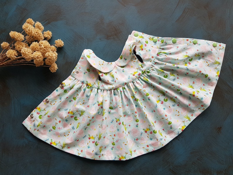 Puede incluir: Un vestido de beb&eacute; azul claro con cuello blanco y estampado floral de flores rosas, hojas verdes y detalles amarillos. El vestido tiene una falda fruncida y se abrocha con botones.